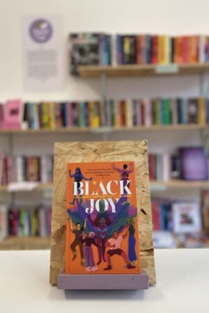Cymraeg: Copi o 'Black Joy' yn sefyll ar stondin llyfrau, tu blaen silffoedd o lyfrau yn y cefndir. English: A copy of 'Black Joy' sits on a stand in front of multiple shelves of other books.