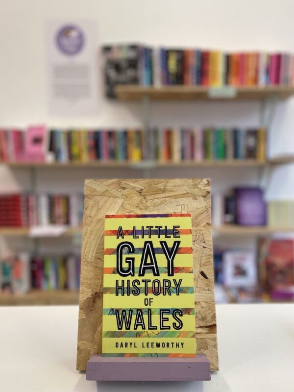 A copy of 'A Little Gay History of Wales' sits on a stand in front of multiple shelves of other books.