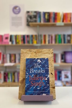 Cymraeg: Copi o 'The Breaks' yn sefyll ar stondin llyfrau, tu blaen silffoedd o lyfrau yn y cefndir. English: A copy of 'The Breaks' sits on a stand in front of multiple shelves of other books.
