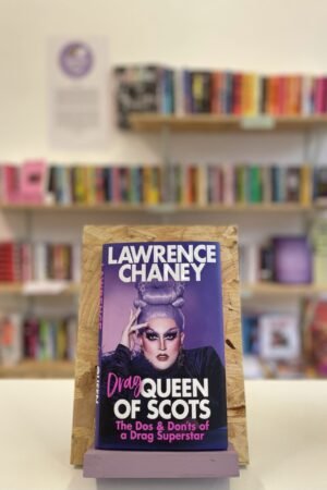 Cymraeg: Copi o 'Drag Queen of Scots' yn sefyll ar stondin llyfrau, tu blaen silffoedd o lyfrau yn y cefndir. | English: A copy of 'Drag Queen of Scots' sits on a stand in front of multiple shelves of other books.