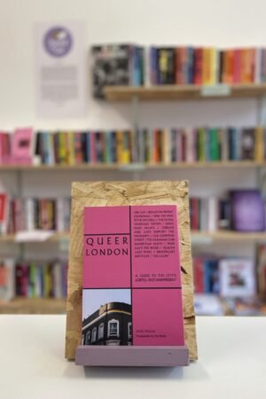 Cymraeg: Copi o 'Queer London' yn sefyll ar stondin llyfrau, tu blaen silffoedd o lyfrau yn y cefndir. | English: A copy of 'Queer London' sits on a stand in front of multiple shelves of other books.