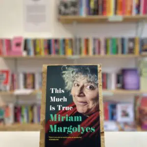 Cymraeg: Copi o 'This Much Is True' yn sefyll ar stondin llyfrau, tu blaen silffoedd o lyfrau yn y cefndir. | English: A copy of 'This Much Is True' sits on a stand in front of multiple shelves of other books.