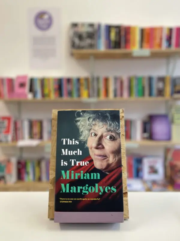 Cymraeg: Copi o 'This Much Is True' yn sefyll ar stondin llyfrau, tu blaen silffoedd o lyfrau yn y cefndir. | English: A copy of 'This Much Is True' sits on a stand in front of multiple shelves of other books.