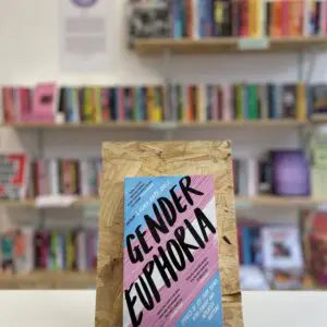 Cymraeg: Copi o 'Gender Euphoria' yn sefyll ar stondin llyfrau, tu blaen silffoedd o lyfrau yn y cefndir. | English: A copy of 'Gender Euphoria' sits on a stand in front of multiple shelves of other books.