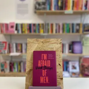 Cymraeg: Copi o 'I'm Afraid of Men' yn sefyll ar stondin llyfrau, tu blaen silffoedd o lyfrau yn y cefndir. | English: A copy of 'I'm Afraid of Men' sits on a stand in front of multiple shelves of other books.