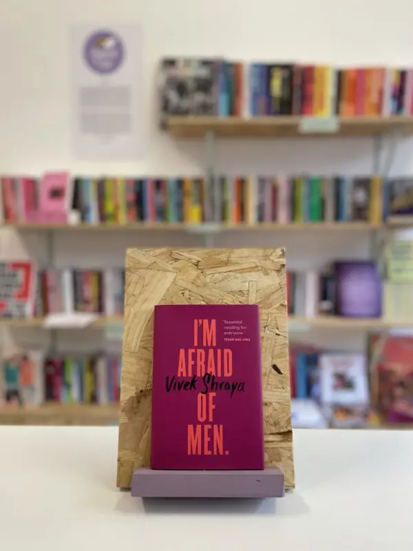 Cymraeg: Copi o 'I'm Afraid of Men' yn sefyll ar stondin llyfrau, tu blaen silffoedd o lyfrau yn y cefndir. | English: A copy of 'I'm Afraid of Men' sits on a stand in front of multiple shelves of other books.