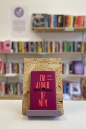 Cymraeg: Copi o 'I'm Afraid of Men' yn sefyll ar stondin llyfrau, tu blaen silffoedd o lyfrau yn y cefndir. | English: A copy of 'I'm Afraid of Men' sits on a stand in front of multiple shelves of other books.