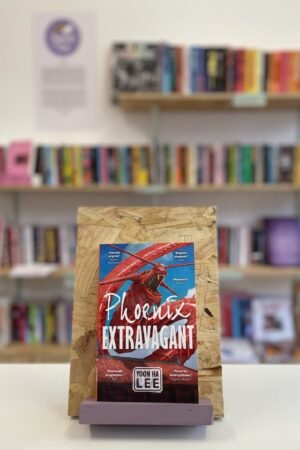 Cymraeg: Copi o 'Phoenix Extravagant' yn sefyll ar stondin llyfrau, tu blaen silffoedd o lyfrau yn y cefndir. | English: A copy of 'Phoenix Extravagant' sits on a stand in front of multiple shelves of other books.