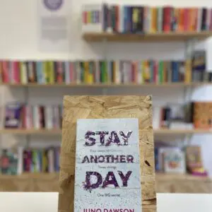Cymraeg: Copi o 'Stay Another Day' yn sefyll ar stondin llyfrau, tu blaen silffoedd o lyfrau yn y cefndir. | English: A copy of 'Stay Another Day' sits on a stand in front of multiple shelves of other books.