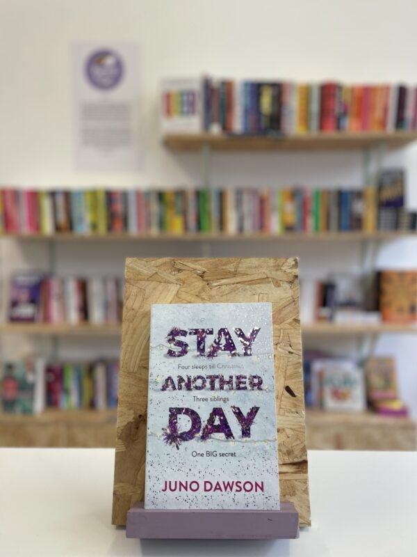 Cymraeg: Copi o 'Stay Another Day' yn sefyll ar stondin llyfrau, tu blaen silffoedd o lyfrau yn y cefndir. | English: A copy of 'Stay Another Day' sits on a stand in front of multiple shelves of other books.
