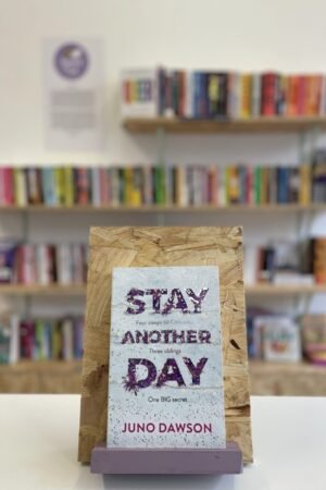 Cymraeg: Copi o 'Stay Another Day' yn sefyll ar stondin llyfrau, tu blaen silffoedd o lyfrau yn y cefndir. | English: A copy of 'Stay Another Day' sits on a stand in front of multiple shelves of other books.
