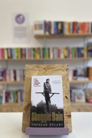 Cymraeg: Copi o 'Shuggie Bain' yn sefyll ar stondin llyfrau, tu blaen silffoedd o lyfrau yn y cefndir. | English: A copy of 'Shuggie Bain' sits on a stand in front of multiple shelves of other books.
