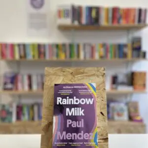 Cymraeg: Copi o 'Rainbow Milk' yn sefyll ar stondin llyfrau, tu blaen silffoedd o lyfrau yn y cefndir. | English: A copy of 'Rainbow Milk' sits on a stand in front of multiple shelves of other books.
