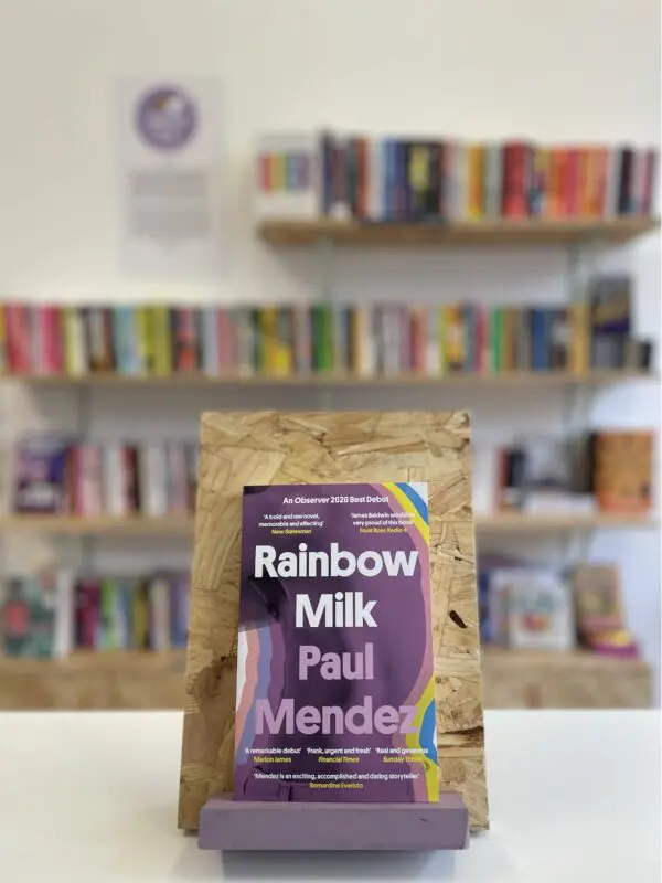 Cymraeg: Copi o 'Rainbow Milk' yn sefyll ar stondin llyfrau, tu blaen silffoedd o lyfrau yn y cefndir. | English: A copy of 'Rainbow Milk' sits on a stand in front of multiple shelves of other books.