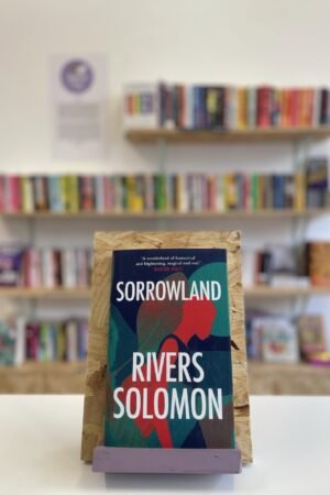 Cymraeg: Copi o 'Sorrowland' yn sefyll ar stondin llyfrau, tu blaen silffoedd o lyfrau yn y cefndir. | English: A copy of 'Sorrowland' sits on a stand in front of multiple shelves of other books.