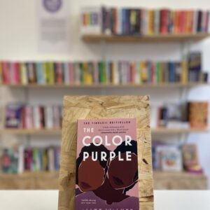 Cymraeg: Copi o 'The Color Purple' yn sefyll ar stondin llyfrau, tu blaen silffoedd o lyfrau yn y cefndir. | English: A copy of 'The Color Purple' sits on a stand in front of multiple shelves of other books.