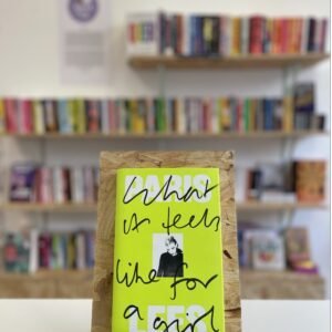 Cymraeg: Copi o 'What It Feels Like to Be a Girl' yn sefyll ar stondin llyfrau, tu blaen silffoedd o lyfrau yn y cefndir. | English: A copy of 'What It Feels Like For a Girl' sits on a stand in front of multiple shelves of other books.