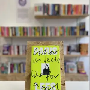 Cymraeg: Copi o 'What It Feels Like to Be a Girl' yn sefyll ar stondin llyfrau, tu blaen silffoedd o lyfrau yn y cefndir. | English: A copy of 'What It Feels Like For a Girl' sits on a stand in front of multiple shelves of other books.