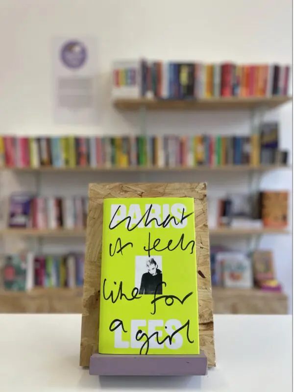 Cymraeg: Copi o 'What It Feels Like to Be a Girl' yn sefyll ar stondin llyfrau, tu blaen silffoedd o lyfrau yn y cefndir. | English: A copy of 'What It Feels Like For a Girl' sits on a stand in front of multiple shelves of other books.