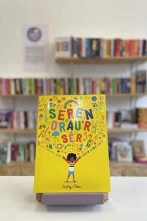 Cymraeg: Copi o 'Seren Orau'r Sêr' yn sefyll ar stondin llyfrau, tu blaen silffoedd o lyfrau yn y cefndir. | English: A copy of 'Seren Orau'r Sêr' sits on a stand in front of multiple shelves of other books.