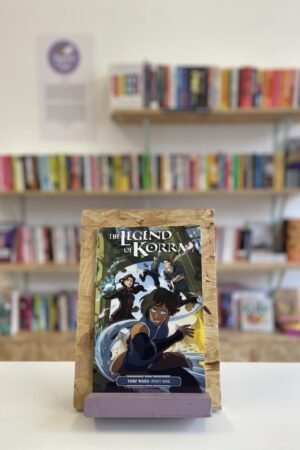Cymraeg: Copi o 'The Legend of Korra: Turf Wars, Part One' yn sefyll ar stondin llyfrau, tu blaen silffoedd o lyfrau yn y cefndir. | English: A copy of 'The Legend of Korra: Turf Wars, Part One' sits on a stand in front of multiple shelves of other books.