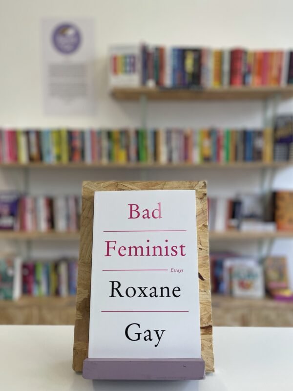 Cymraeg: Copi o 'The Color Purple' yn sefyll ar stondin llyfrau, tu blaen silffoedd o lyfrau yn y cefndir. | English: A copy of 'The Color Purple' sits on a stand in front of multiple shelves of other books.