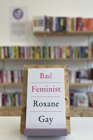Cymraeg: Copi o 'The Color Purple' yn sefyll ar stondin llyfrau, tu blaen silffoedd o lyfrau yn y cefndir. | English: A copy of 'The Color Purple' sits on a stand in front of multiple shelves of other books.