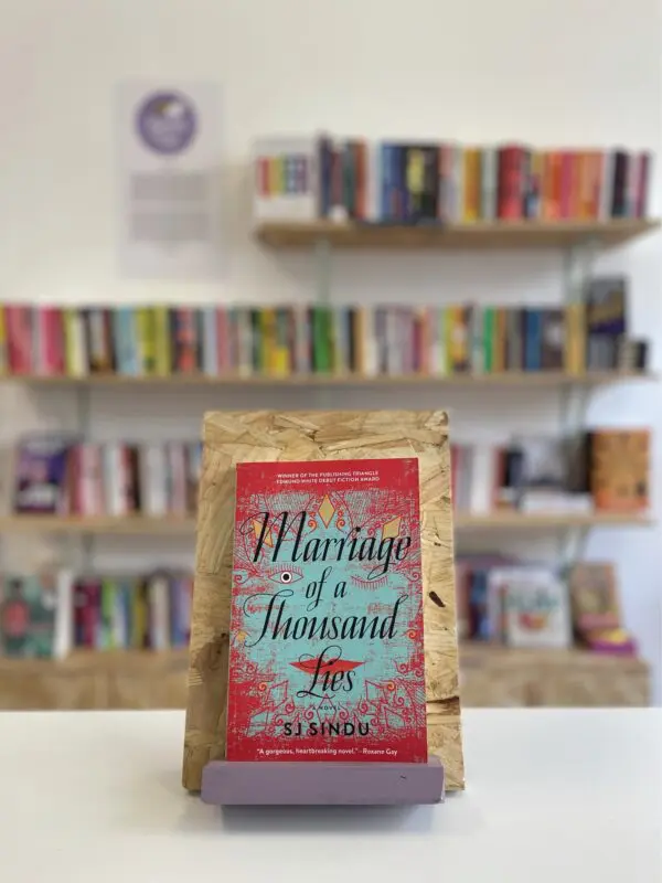 A copy of 'Marriage of a Thousand Lies' sits on a stand in front of multiple shelves of other books.