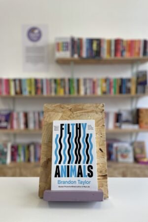 Cymraeg: Copi o 'Filthy Animals' yn sefyll ar stondin llyfrau, tu blaen silffoedd o lyfrau yn y cefndir. | English: A copy of 'Filthy Animals' sits on a stand in front of multiple shelves of other books.