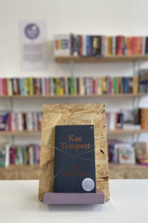 Cymraeg: Copi o 'On Connection' yn sefyll ar stondin llyfrau, tu blaen silffoedd o lyfrau yn y cefndir. | English: A copy of 'On Connection' sits on a stand in front of multiple shelves of other books.