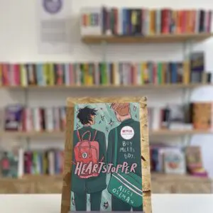 A copy of 'Heartstopper vol. 1' sits on a stand in front of multiple shelves of other books.