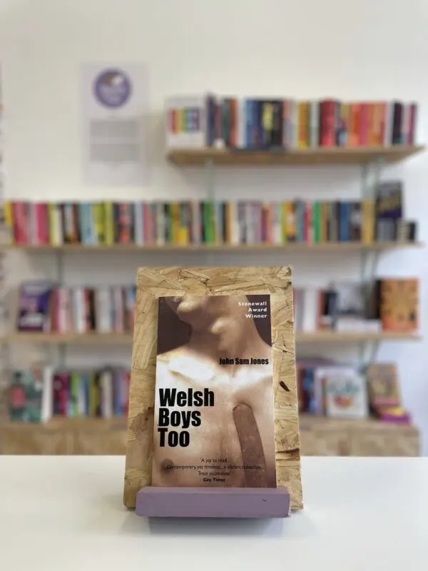 Cymraeg: Copi o 'Welsh Boys Too' yn sefyll ar stondin llyfrau, tu blaen silffoedd o lyfrau yn y cefndir. | English: A copy of 'Welsh Boys Too' sits on a stand in front of multiple shelves of other books.