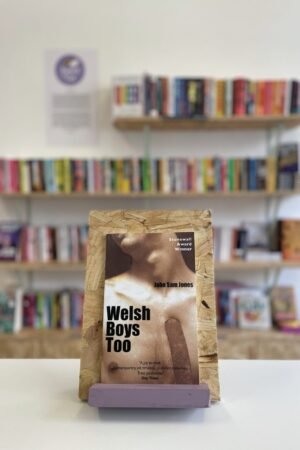 Cymraeg: Copi o 'Welsh Boys Too' yn sefyll ar stondin llyfrau, tu blaen silffoedd o lyfrau yn y cefndir. | English: A copy of 'Welsh Boys Too' sits on a stand in front of multiple shelves of other books.