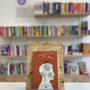 Cymraeg: Copi o 'Y Pump: Tim' yn sefyll ar stondin llyfrau, tu blaen silffoedd o lyfrau yn y cefndir. | English: A copy of 'Y Pump: Tim' sits on a stand in front of multiple shelves of other books.