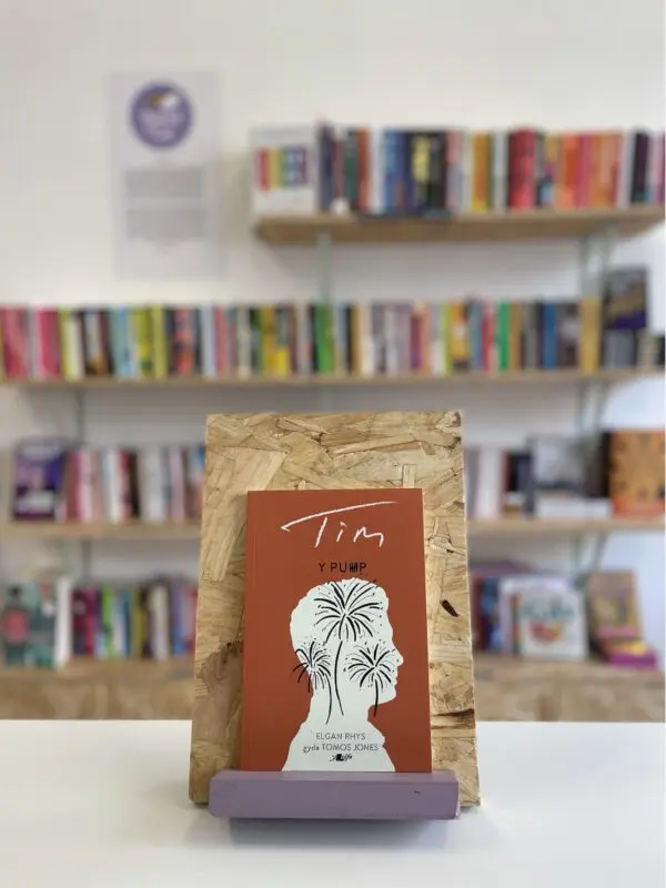 Cymraeg: Copi o 'Y Pump: Tim' yn sefyll ar stondin llyfrau, tu blaen silffoedd o lyfrau yn y cefndir. | English: A copy of 'Y Pump: Tim' sits on a stand in front of multiple shelves of other books.