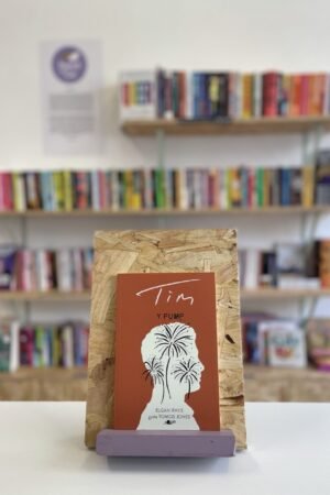 Cymraeg: Copi o 'Y Pump: Tim' yn sefyll ar stondin llyfrau, tu blaen silffoedd o lyfrau yn y cefndir. | English: A copy of 'Y Pump: Tim' sits on a stand in front of multiple shelves of other books.