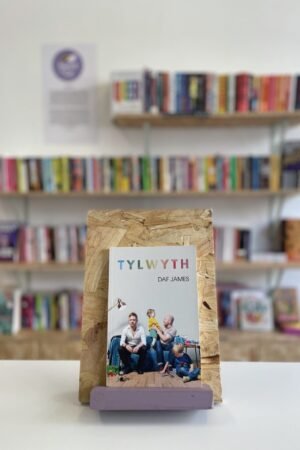 Cymraeg: Copi o 'Tylwyth' yn sefyll ar stondin llyfrau, tu blaen silffoedd o lyfrau yn y cefndir. | English: A copy of 'Tylwyth' sits on a stand in front of multiple shelves of other books.