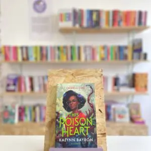 Cymraeg: Copi o 'The Poison Heart' yn sefyll ar stondin llyfrau, tu blaen silffoedd o lyfrau yn y cefndir. | English: A copy of 'The Poison Heart' sits on a stand in front of multiple shelves of other books.