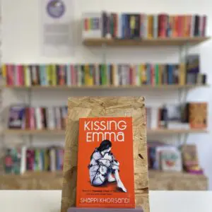 Cymraeg: Copi o 'Kissing Emma' yn sefyll ar stondin llyfrau, tu blaen silffoedd o lyfrau yn y cefndir. | English: A copy of 'Kissing Emma' sits on a stand in front of multiple shelves of other books.