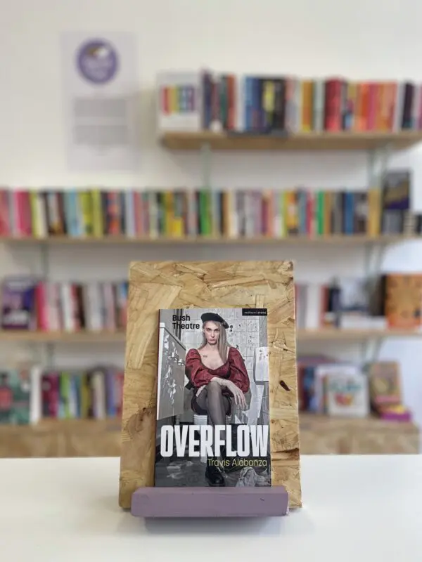 Cymraeg: Copi o 'Overflow' yn sefyll ar stondin llyfrau, tu blaen silffoedd o lyfrau yn y cefndir. English: A copy of 'Overflow' sitson a stand in front of multiple shelves of other books.