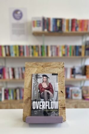 Cymraeg: Copi o 'Overflow' yn sefyll ar stondin llyfrau, tu blaen silffoedd o lyfrau yn y cefndir. English: A copy of 'Overflow' sitson a stand in front of multiple shelves of other books.
