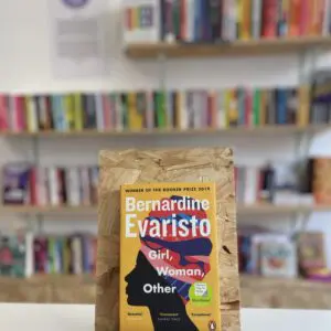 Cymraeg: Copi o 'Girl, Woman, Other' yn sefyll ar stondin llyfrau, tu blaen silffoedd o lyfrau yn y cefndir. | English: A copy of 'Girl, Woman, Other' sits on a stand in front of multiple shelves of other books.