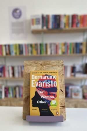 Cymraeg: Copi o 'Girl, Woman, Other' yn sefyll ar stondin llyfrau, tu blaen silffoedd o lyfrau yn y cefndir. | English: A copy of 'Girl, Woman, Other' sits on a stand in front of multiple shelves of other books.