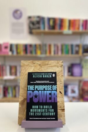 Cymraeg: Copi o 'The Purpose of Power' yn sefyll ar stondin llyfrau, tu blaen silffoedd o lyfrau yn y cefndir. | English: A copy of 'The Purpose of Power' sits on a stand in front of multiple shelves of other books.