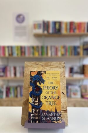Cymraeg: Copi o 'The Priory of the Orange Tree' yn sefyll ar stondin llyfrau, tu blaen silffoedd o lyfrau yn y cefndir. | English: A copy of 'The Priory of the Orange Tree' sits on a stand in front of multiple shelves of other books.