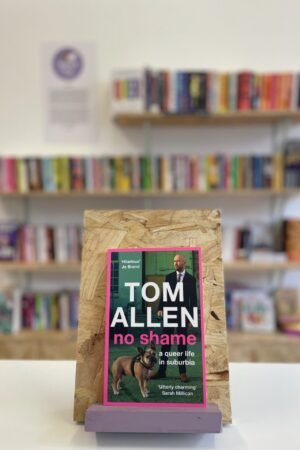 Cymraeg: Copi o 'No Shame' yn sefyll ar stondin llyfrau, tu blaen silffoedd o lyfrau yn y cefndir. | English: A copy of 'No Shame' sits on a stand in front of multiple shelves of other books.