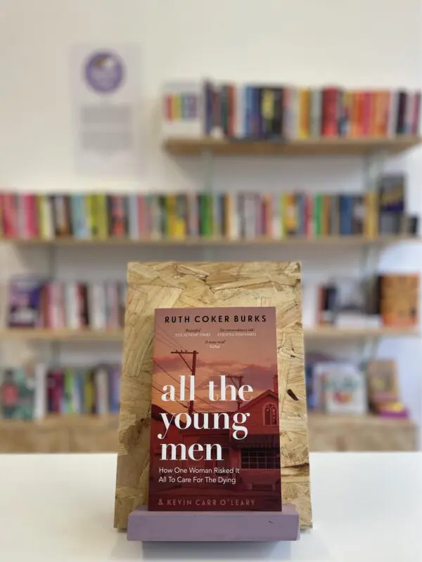 Cymraeg: Copi o 'All the Young Men' yn sefyll ar stondin llyfrau, tu blaen silffoedd o lyfrau yn y cefndir. | English: A copy of 'All the Young Men' sits on a stand in front of multiple shelves of other books.