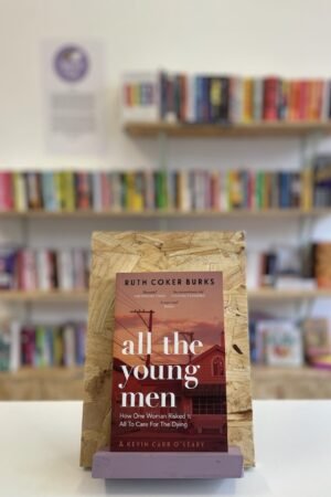 Cymraeg: Copi o 'All the Young Men' yn sefyll ar stondin llyfrau, tu blaen silffoedd o lyfrau yn y cefndir. | English: A copy of 'All the Young Men' sits on a stand in front of multiple shelves of other books.