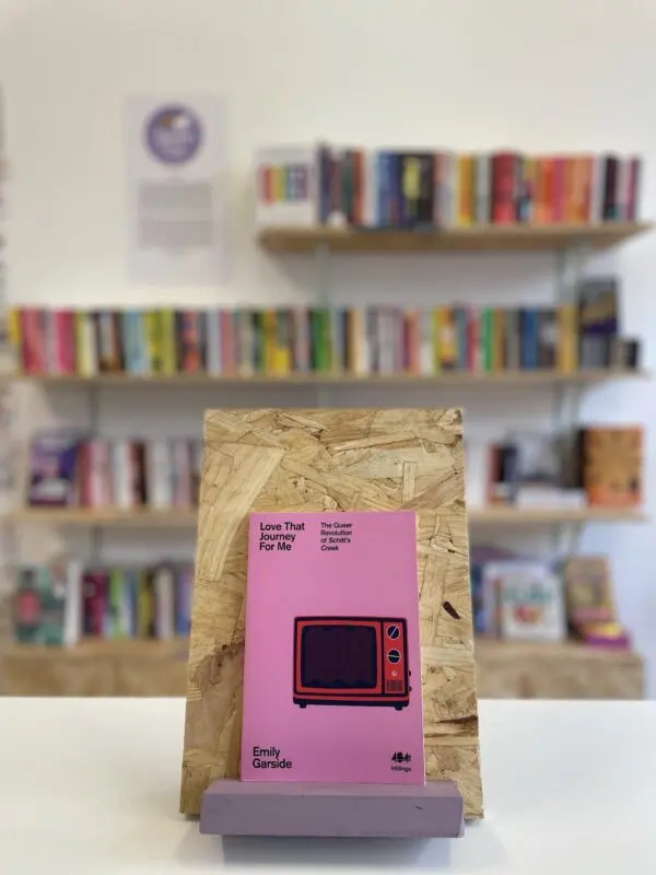Cymraeg: Copi o 'Love That Journey For Me' yn sefyll ar stondin llyfrau, tu blaen silffoedd o lyfrau yn y cefndir. English: A copy of 'Love That Journey For Me' sits on a stand in front of multiple shelves of other books.