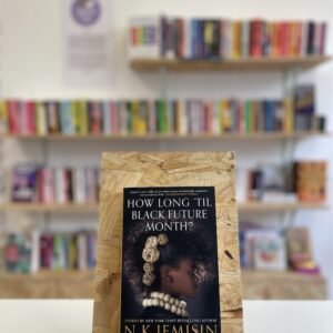 Cymraeg: Copi o 'How Long 'Til Black Future Month?' yn sefyll ar stondin llyfrau, tu blaen silffoedd o lyfrau yn y cefndir. English: A copy of 'How Long 'Til Black Future Month?' sits on a stand in front of multiple shelves of other books.